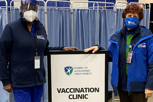 two women wearing face mask in front of a sign with the Roanoke City and Alleghany Health Districts logo and "Vaccination Clinic" printed on it.