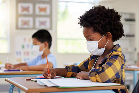 Niño con mascarilla en el aula sentado en el escritorio trabajando en el trabajo de clase.