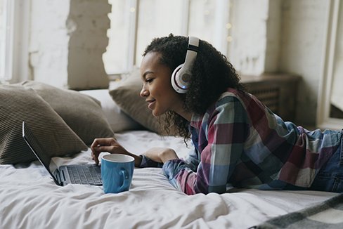 Girl relaxing on couch with music and coffee while browsing on laptop.