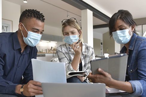 group of 3 people gathered around a laptop while wearing surgical facemasks to protect from COVID-19
