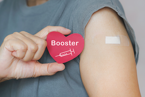 hand holding a small cardboard heart that says "booster" with an image of a syringe next to a bandage on their other arm