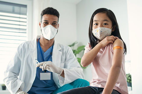 Bác sĩ cầm ống tiêm rỗng và bé gái khoe băng quấn trên cánh tay sau khi được tiêm vắc-xin.