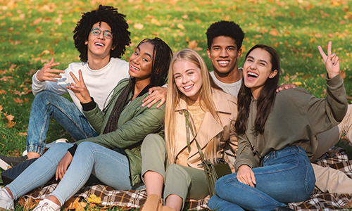 Multiethnic Group of Happy Teenagers Having Fun In Autumn Park, sitting on the ground and posing at camera