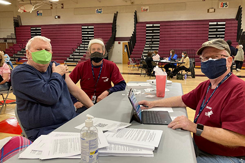 Man who just received COVID-19 vaccination sitting with two men from the Medical Reserve Corps, all wearing surgical masks while one works on a computer