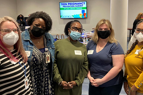 Group of 5 women standing and smiling while wearing surgical masks.