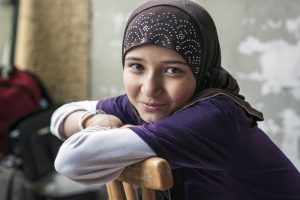 A smiling girl in a headscarf leans over the back of a chair with her arms crossed.