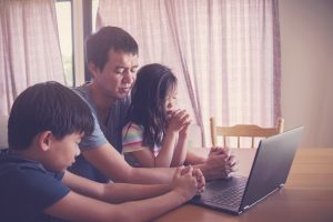 Father and two children praying in front of a computer 
