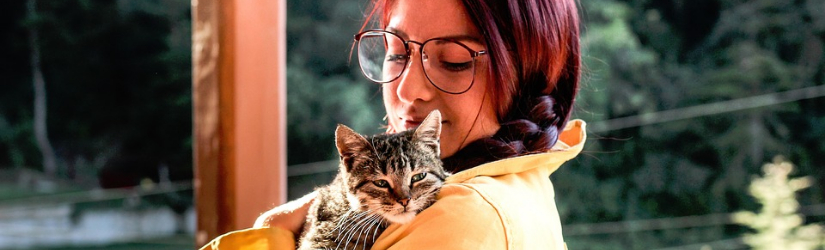 A person with braided red hair, glasses, and a yellow-orange shirt cradles a tabby cat over her shoulder.