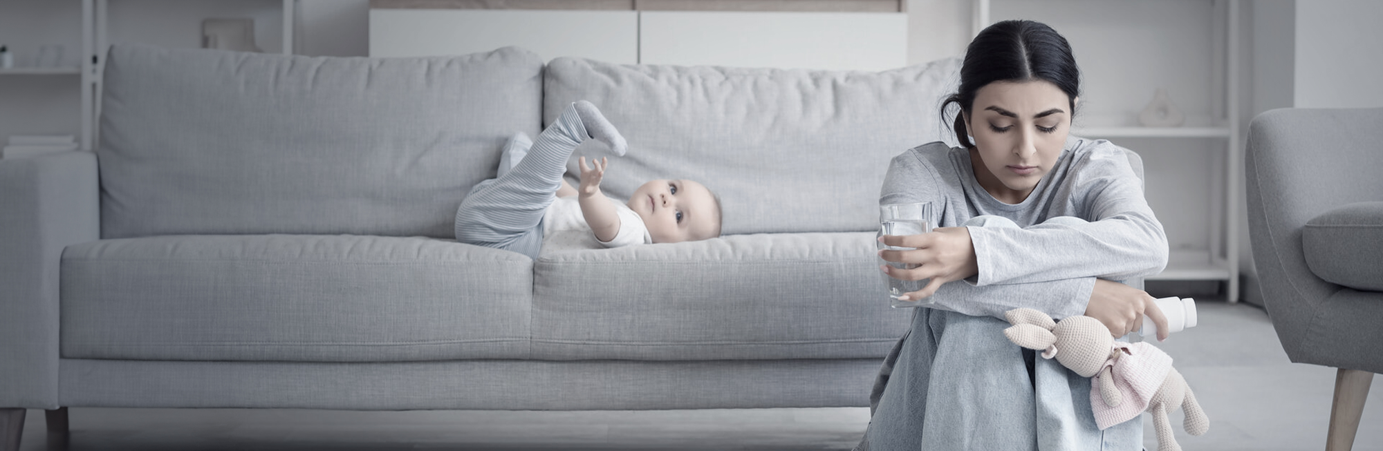 A depressed woman sitting on the ground, with a baby lying on the couch behind her