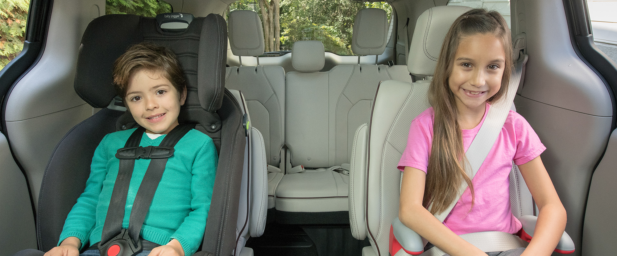 1 child in a car seat next to an older child wearing a seat belt