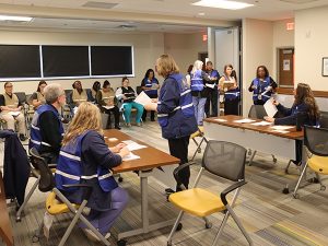 People in tan vests sit in chairs while people in blue vests are sitting and standing.