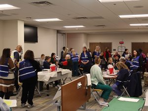 A room full of people sitting at tables and standing, wearing vests.
