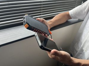 한 사람이 창문 옆에 서서 한 손에는 태양열 패널이 달린 충전 장치를 들고 다른 한 손에는 모바일 기기를 들고 있습니다.